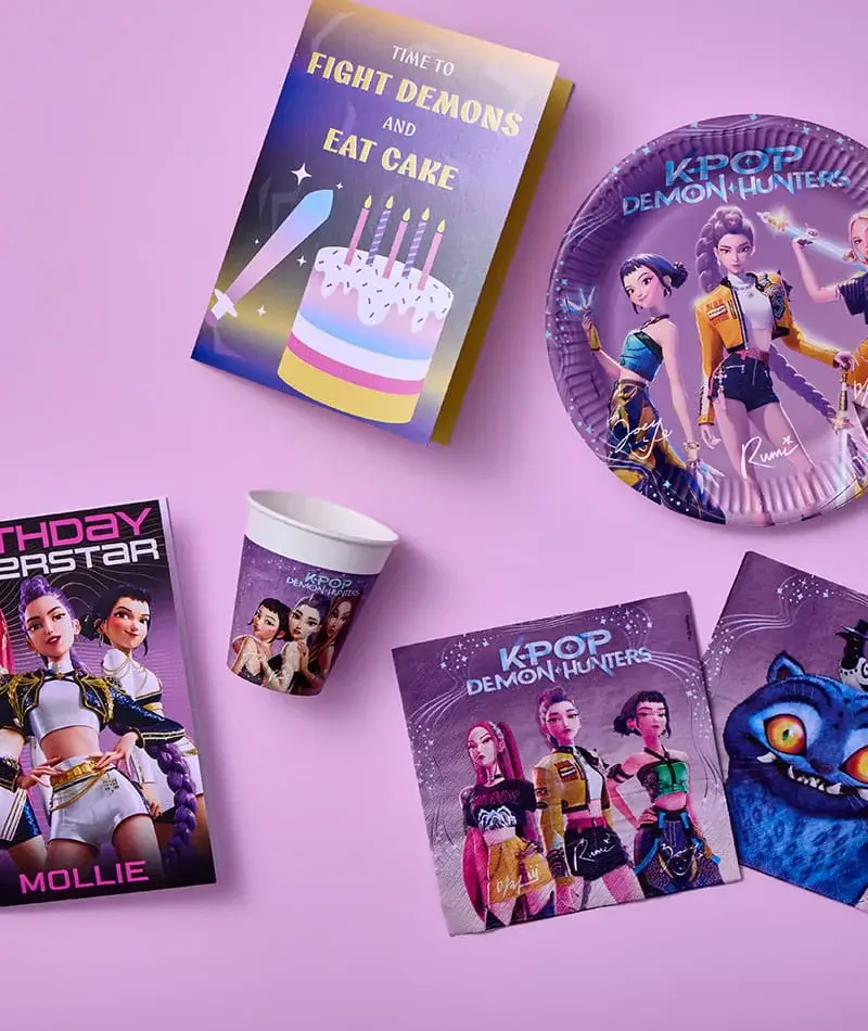 K‑pop themed party supplies arranged on a pink surface, including illustrated plates, cups, cards, and decorations featuring fantasy characters and vibrant colors.
